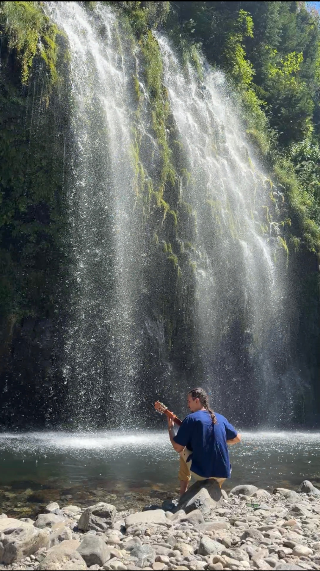 Cielo playing guitar on a rock at the foot of a waterfall